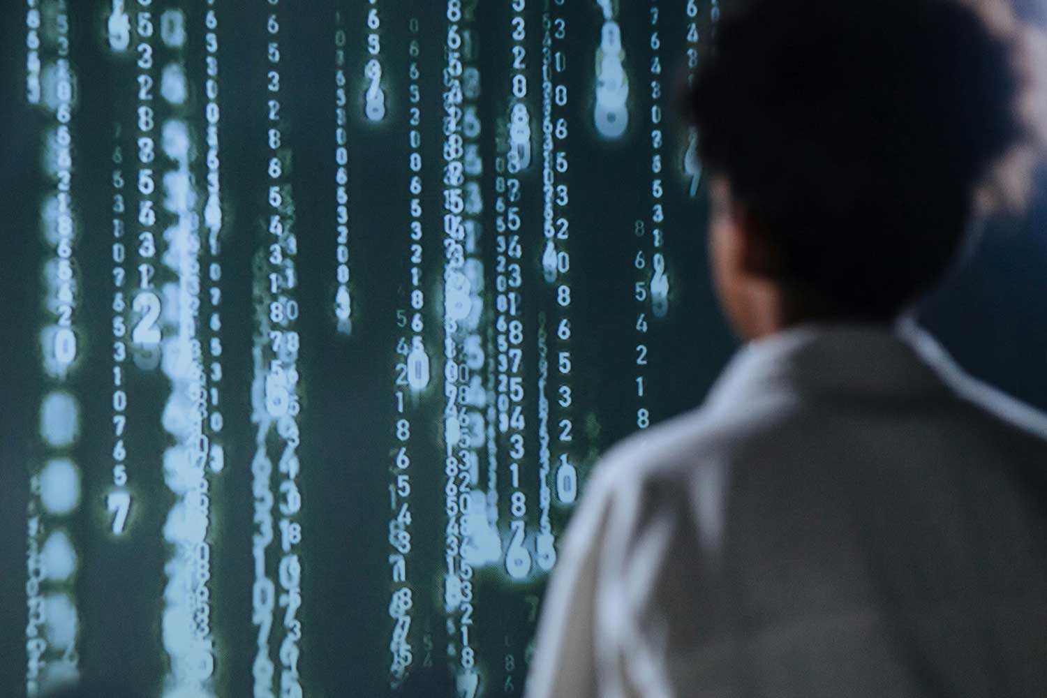 Person viewed from behind a screen with vertical rows of illuminated numbers and binary code, similar to a matrix display.