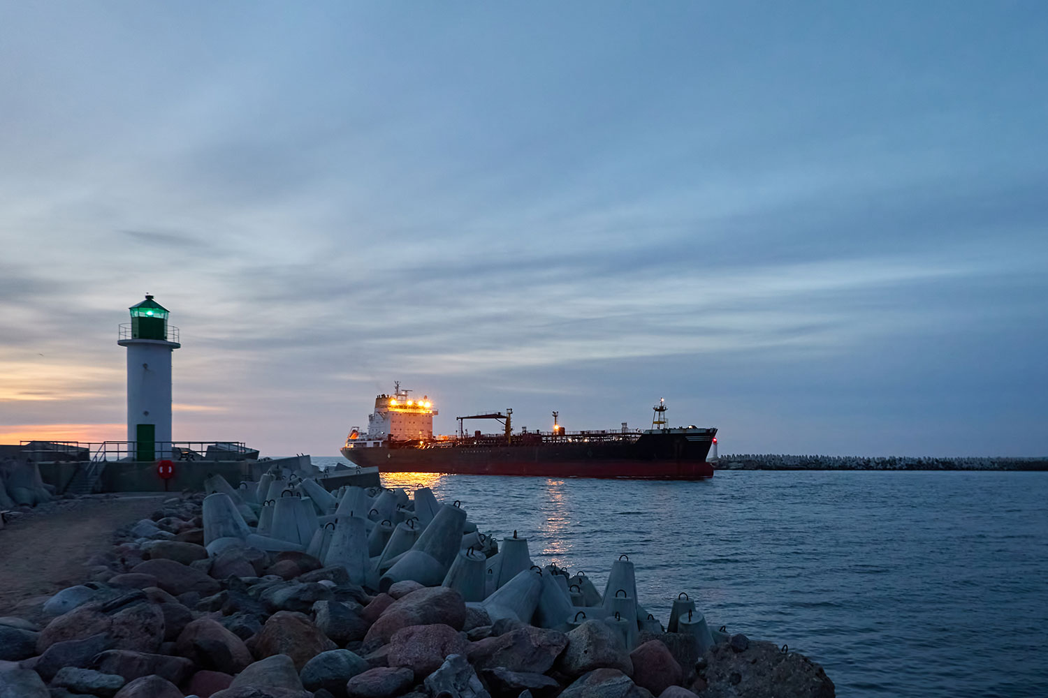 Großes Frachtschiff bei der Ankunft im Frachthafen bei Sonnenuntergang. Wellenbrecher, Promenade zum Leuchtturm. Seelandschaft in Ventspils, Lettland. | Quelle: AdobeStock by Aastels.