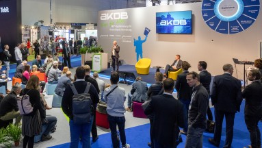 AKDB stand at the Smart Country Convention 2023. In the background a man on stage, in the foreground listeners from behind.