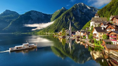 Landschaftsbild Österreich aus den Bergen, mit einem Bergdorf rechts, einem See links und einem Schiff auf dem Wasser 