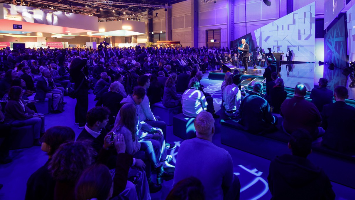 Stage of the Smart Country Convention with audience in the foreground, Ralf Wintergerst is on stage on the right, trade fair stands are shown on the left