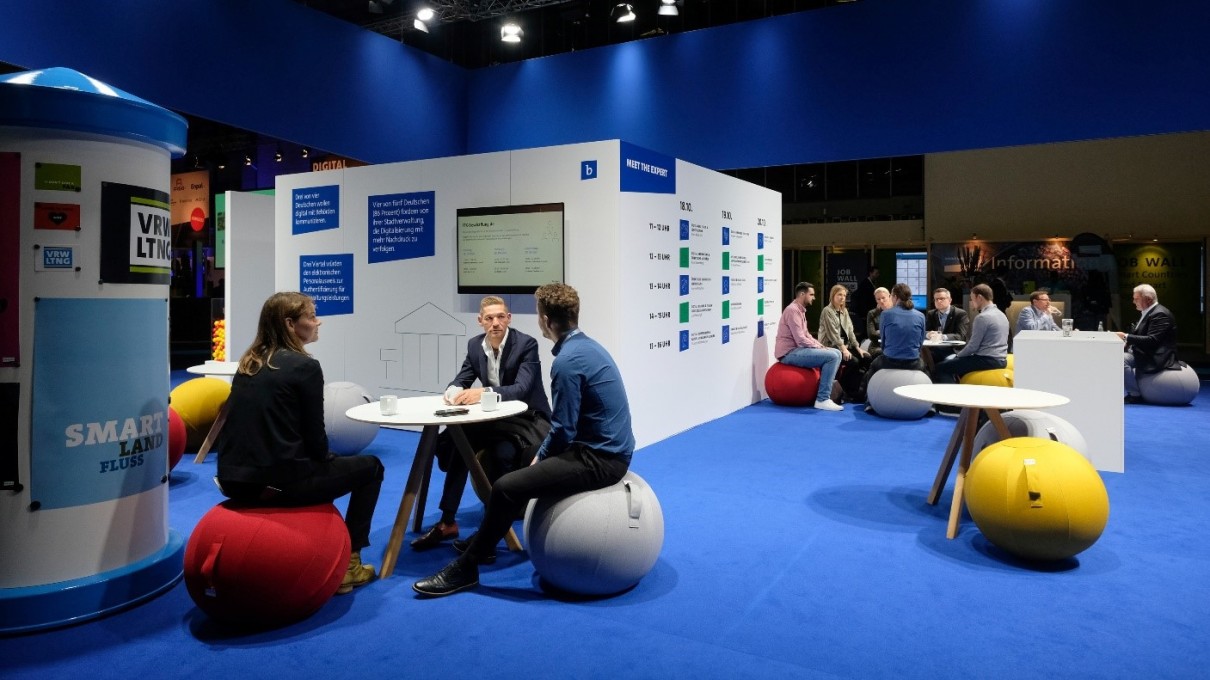 Der Messestand vom Bitkom auf der Smart Country Convention. Im Vordergrund Menschen, die auf Bällen sitzen und sich unterhalten. Links eine Litfaßsäule mit Postern, im Hintergrund weitere Personen