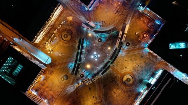 Aerial view at night, large, illuminated town square has a symmetrical structure with curved street lines and tram tracks.