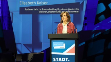 Elisabeth Kaiser, Parliamentary State Secretary at the BMWSB, stands behind a lectern on the stage and gives a speech