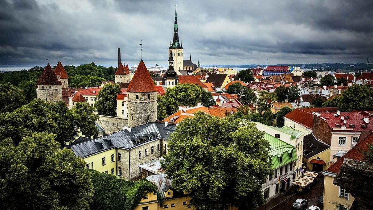 Blick auf die Altstadt von Tallinn, Estland, mit mittelalterlichen Türmen, roten Dächern und grünen Bäumen, die unter einem dramatisch bewölkten Himmel hervorragen.