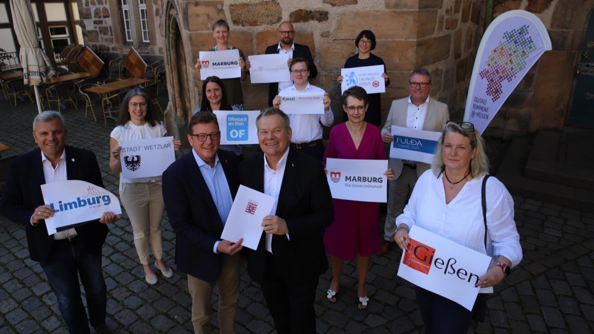 9 municipalities and districts receive a grant from State Secretary Sauer. The participants hold up signs with the logos of the 9 towns and districts. 