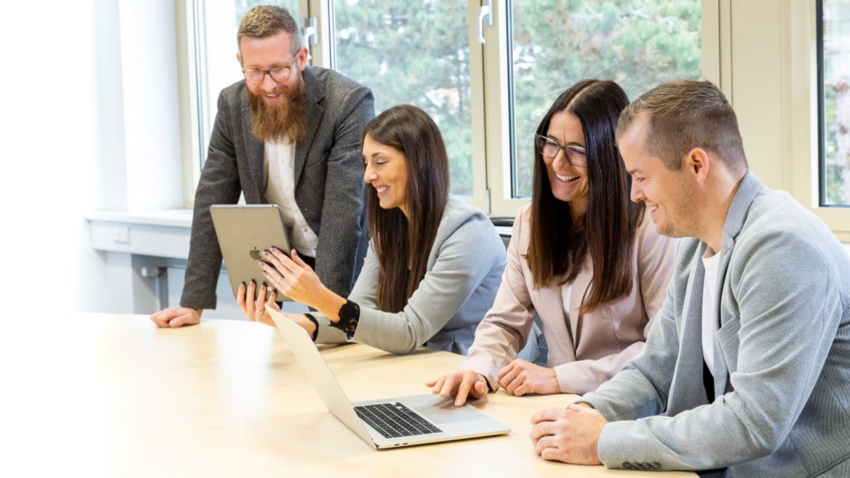 4 people are sitting at a desk in an office. 2 women explain something to 2 men at a computer