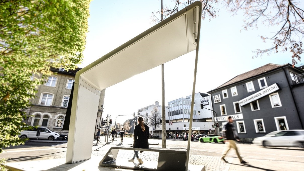 Smarte Bank in Heidenheim auf der eine Frau sitzt im Vordergrund, im Hintergrund ein Blick in die Stadt mit Gebäuden, Menschen und Autos 