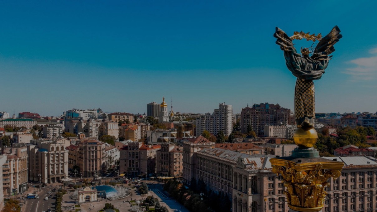 Panoramablick auf den Freiheitsplatz in Kiew, Ukraine, mit dem Unabhängigkeitsdenkmal im Vordergrund, umgeben von historischen Gebäuden und moderner Architektur unter einem klaren blauen Himmel.