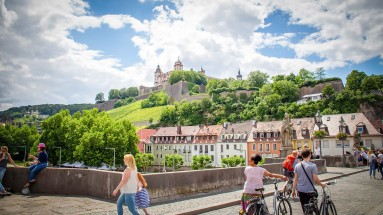 Stadtansicht von Würzburg mit Menschen im Vordergrund, einer Häuserreihe in der Mitte und der Würzburger Festung im Hintergrund 