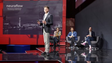 Junger Mann präsentiert auf der Bühne des Smart Country Startup Award 2024 vor einer Leinwand mit dem Schriftzug 'neuraflow', drei Personen sitzen im Hintergrund auf Stühlen. Junger Mann präsentiert auf der Bühne des Smart Country Startup Award 2024 vor einer Leinwand mit dem Schriftzug 'neuraflow', drei Personen sitzen im Hintergrund auf Stühlen.
