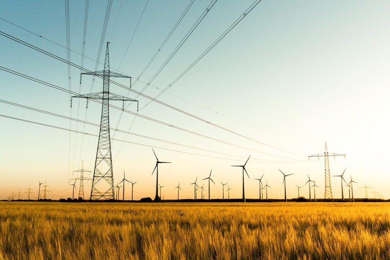 Strommasten, Kornfeld und Windräder im herbstlichen Gegenlicht. Symbolbild einer zukunftstauglichen Infrastruktur für die Energieverteilung – das Smart Grid.